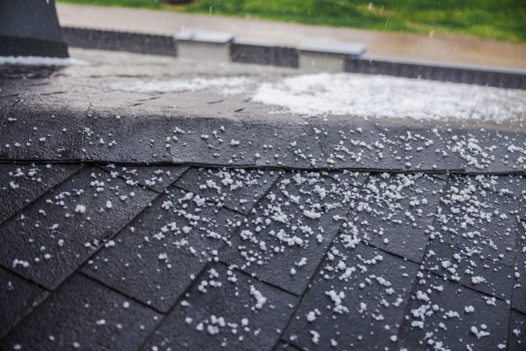 Small hailstones falling and covering a dark shingled roof during a rainstorm.